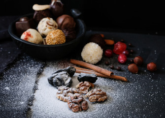 A variety of truffles in a black clay pile, walnut kernels and prunes under powdered sugar, cinnamon sticks and fresh cranberries