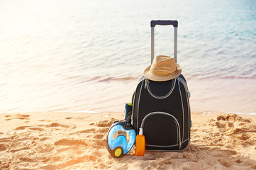 Suitcase and hat, sunscreen with a mask. The tropical sea, beach in the background. The concept of summer recreation travel and cruise traffic