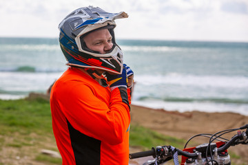 Motorcyclist in a protective suit stands next to motorbike in front of the sea