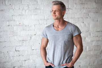Fototapeta premium Young sexy men bodybuilder athlete studio portrait in loft on the background of white brick wall. Handsome guy model in grey T-shirt