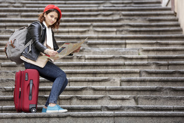 Giovane  con basco rosso seduta nel sopra il suo trolley rosso in una scalinata guarda una mappa in...
