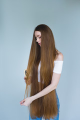 Concept of hair loss. Close up portrait of unhappy sad stressed young woman with long dry brown hair, she is looking at the brush in hand, isolated on grey background.