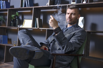 elegant man in suit sits in chair and smokes pipe. handsome stylish rich brutal man in luxurious...