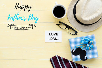 Happy fathers day concept. Red tie, glasses, hat, mustache, gift box with Love DAD text on black heart tag on bright yellow pastel wooden table background.