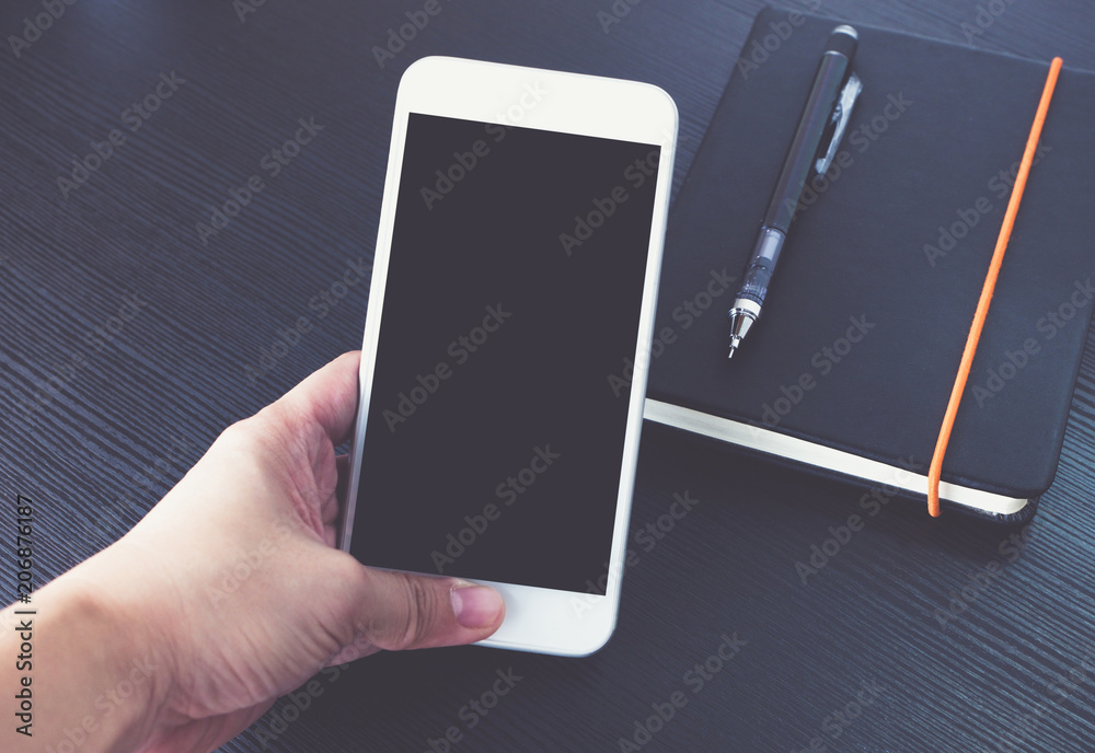 Poster hand holding blank screen of mobile phone over black notebook on black wooden office desk, mock up f