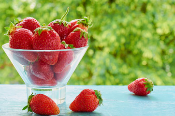 Fresh strawberries in a glass bowl on the blue kitchen table on the blurred nature background. Tasty vegan summer dessert. Ingredients for healthy smoothie. Close up view