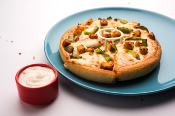 Paneer Pizza is an indian version of Italian dish topped with Cottage Cheese, served in a plate with white sauce. selective focus