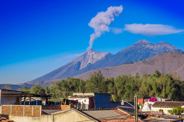 Beautiful landscape of huge mountain in process of aruption with a column of ash, view from the rooftops of the building in Antigua city in gorgeous sunny day and blue sky