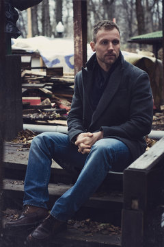 Handsome Young Man In Outerwear And Scarf Posing Outside Of Wooden House Looking At Camera.