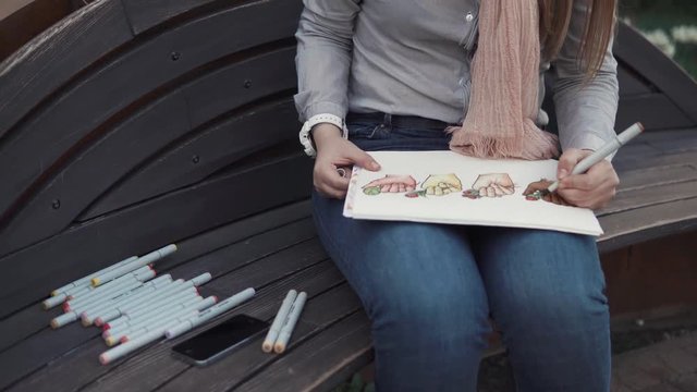 close up shot of the artist's hands, the lady takes up painting on a bench in the park, next to her laid out markers, she finishes drawing an art object