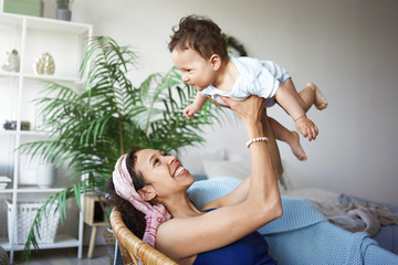 Obraz na płótnie Canvas Happy childhood concept. Indoor shot of casually dressed beautiful young mixed race mom sitting in chair at home and throwing her excited infant daughter up in the air while having fun together