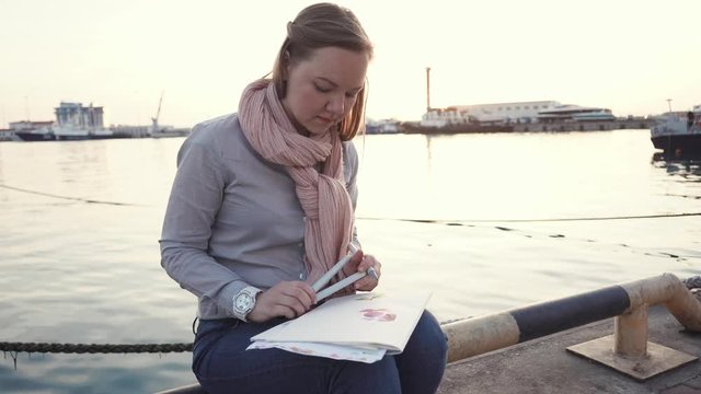 a young woman is engaged in fishing in the port area near the pier, the lady holds the markers and sketch paper in her hands, they are in her thoughts during drawing