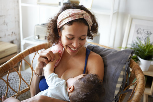 Motherhood, infantry, parenting and childhood concept. Picture of cheerful young brunette Hispanic woman wearing headscarf smiling broadly, nursing her infant child in cozy bedroom interior