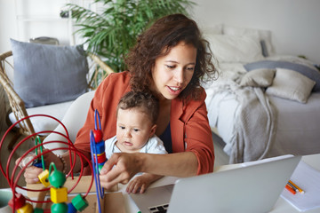Active young mixed race businesswoman multitasking at home, playing with her baby son and checking financial report at the same time, sitting at table in front of open generic portable computer