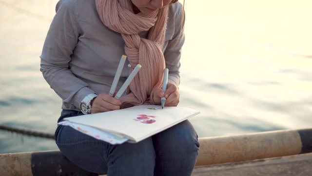 a young woman holds colored pens in her hands and draws flowers, while waiting for her friends, she is near the water in the cool autumn season