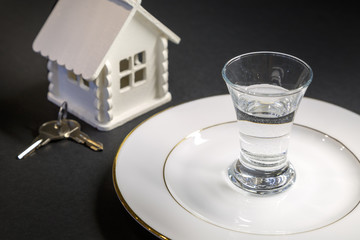 A shot glass of vodka on a white plate against the backdrop of a miniature house and the keys on a black background
