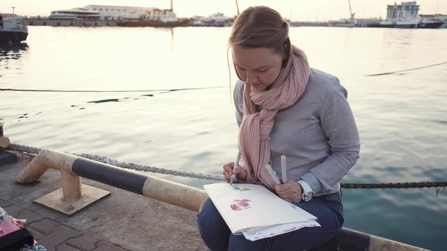 young women who are warmly dressed, finding near the sea or the river, the lady is holding pen markers and is engaged in drawing, the artist is on the planner