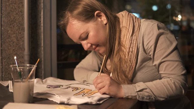 a young and creative woman is engaged in drawing with the help of azure paints, she is driving a tassel on a white T-shirt, to get a fashion picture