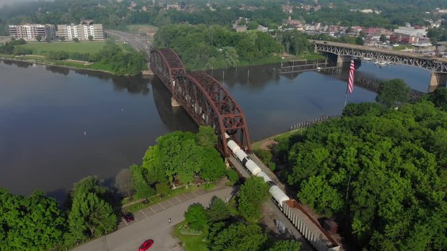A slow reverse morning aerial establishing shot view of a railroad bridge as a freight train passes in the foreground. Tilt up to reveal a small town in the distance. Pittsburgh suburbs.  	