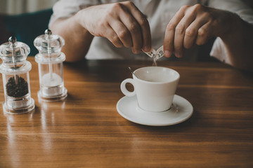 closeup of man's hands pour sugar from a stick into a cup of coffee on a wooden table with salt and pepper