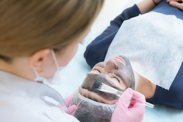 Obraz na płótnie Canvas The cosmetologist in pink gloves with a brush applies a carbon mask for peeling on the face of a young girl in a cosmetology room. The concept of cosmetology services and self-care. The concept of