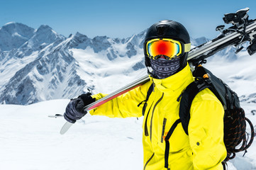 Fototapeta na wymiar A large portrait of a skier in a protective helmet and glasses is a mask and scarf with skis on his shoulder in the snow-capped mountains of the Caucasus. Skiing,