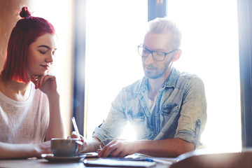 Two friends sitting at cafe table in daylight. Young man in eyeglasses explaining something to his girlfriend, helping her to do homework or sharing his business ideas over cup of coffee