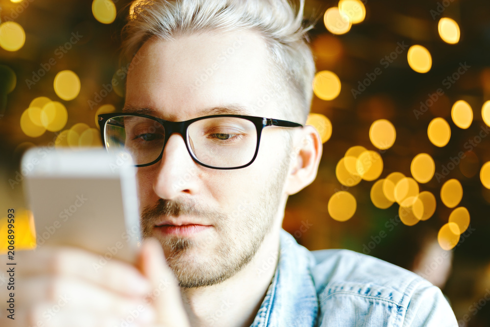 Wall mural portrait of young man in eyeglasses texting on his smartphone on the background of bokeh lights