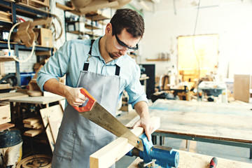 Skillful carpenter sawing a piece of wood placed in workbench vice with hand saw in his workshop