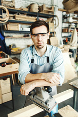 Portrait of confident young woodworker in protective eyeglasses looking at camera while leaning on sanding machine in his workshop