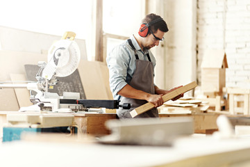 Young carpenter wearing protective eyeglasses and earmuffs preparing to saw wood plank with electric circular saw.