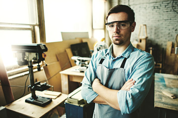 Portrait of confident young carpenter wearing apron and protective eyeglasses posing cross-armed in his woodworking shop.