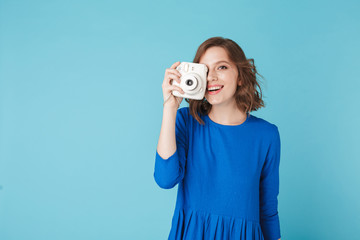 Portrait of young beautiful lady in dress standing with little white camera on over blue...