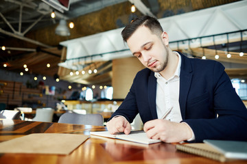 Restaurant manager in shirt and jacket sitting at table and making notes in his personal organizer