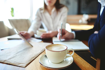 Two defocused business people using tablet computer at coffee meeting, cup of cappucino coffee on foreground