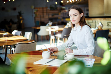 Portrait of young businesswoman looking at camera while working in cafe. Coffee cup, notebooks and documents on her table, pen and tablet computer in her hands