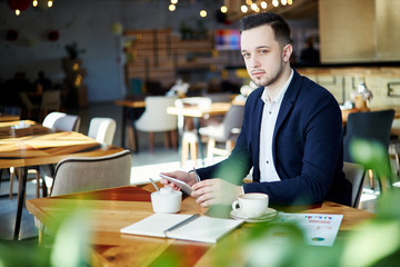 Portrait of confident businessman with stubble beard looking at camera while working in cafe. Coffee cup, notebooks and documents on his table, pen and tablet computer in his hands