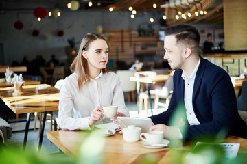 Two young business partners sitting at table in cafe, drinking coffee and discussing work. Businessman holding tablet computer and listening to female colleague
