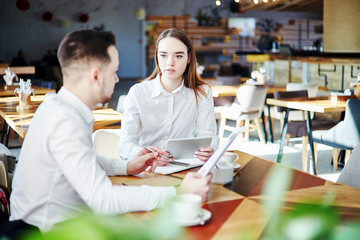 Young business people discussing project at cafe. Businessman with documents in his hands and businesswoman with tablet computer sitting at table and talking