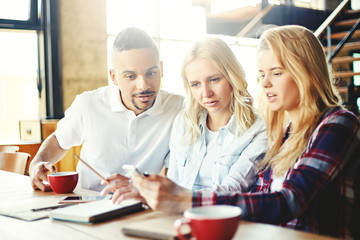 Obraz premium Group of three curious college students sitting at desk and looking at smartphone screen together, using app or browsing pictures