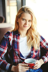 Portrait of pretty blonde woman in checkered shirt drinking coffee in cafe and looking at somebody with interest
