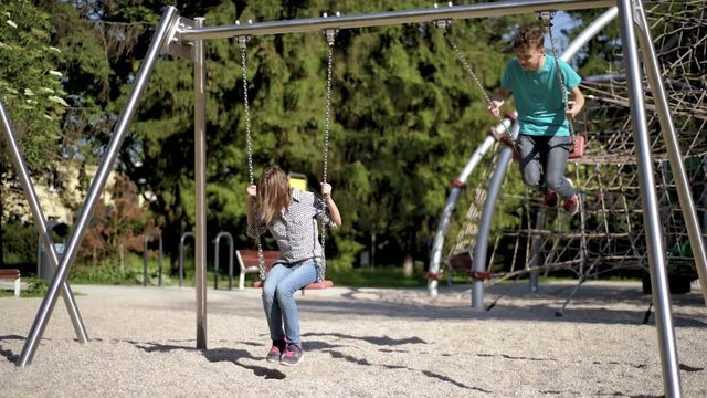 Happy two children ride on a swing at summer park. Cute teen boy and girl swings at playground outdoors.