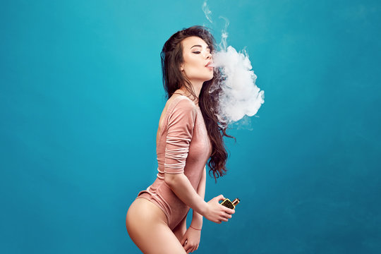 Smiling hot woman in bodysuit standing and vaping on blue background.