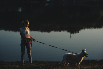 Obraz na płótnie Canvas Side view of a long haired caucasian girl walking in the park around the lake with her white siberian husky at the sunset.