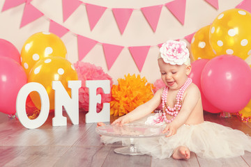 Portrait of cute adorable Caucasian baby girl in tutu tulle skirt celebrating her first birthday. Child kid sitting on floor in studio with pink flags and balloons holding cake stand