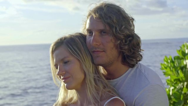 Handheld shot of happy young man sitting on cliff by ocean and hugging his cheerful girlfriend while enjoying summer day on vacation