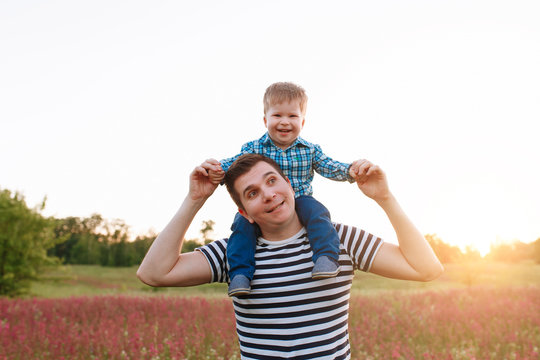 Small boy is sitting on his fathers shoulders and holding dad hands at the sunet in the field during. Copy space for text