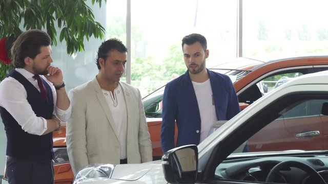Three male friends examining cars for sale at the dealership showroom. Mature Hispanic man and his younger friends shopping for a new automobile. Communication, friendship, consumerism.