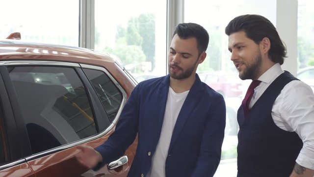 Handsome young man discussing a new automobile for sale with his friend while shopping for a car at the dealership showroom. Male friends choosing automobiles.