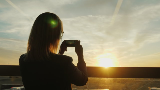 A woman in a business suit takes a photograph of a sunset in a window. In an office building or airport terminal